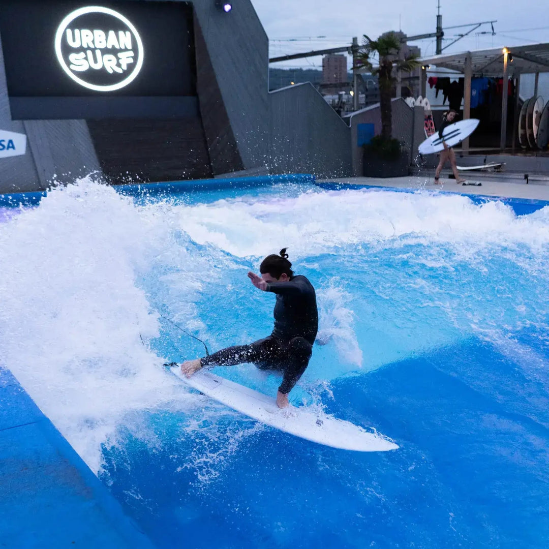 Eine Person im Neoprenanzug surft eine Citywave auf einer künstlichen Indoor-Welle. In der Nähe läuft jemand am Urban Surf-Schild vorbei und trägt ein NOA Surfboards - Pocket Knife 2.0 Board von NOA.