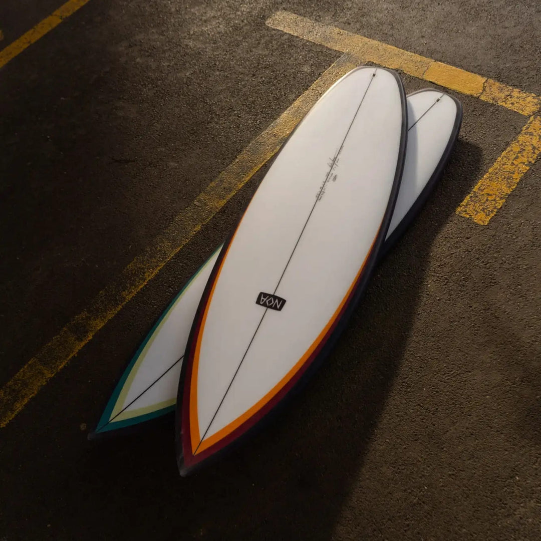 Zwei NOA Surfboards - Pocket Knife 2.0 Boards von NOA liegen auf Asphalt mit gelben Linien. Ihre weißen Tops und spitzen Tips überlappen sich, wobei das nächstgelegene Board in leuchtendem Orange und Rot gehalten ist - ideal für Allround- oder Flusssurfen.