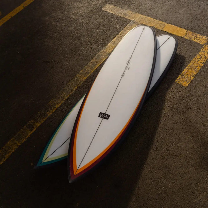 Zwei NOA Surfboards - Pocket Knife 2.0 Boards von NOA liegen auf Asphalt mit gelben Linien. Ihre weißen Tops und spitzen Tips überlappen sich, wobei das nächstgelegene Board in leuchtendem Orange und Rot gehalten ist - ideal für Allround- oder Flusssurfen.