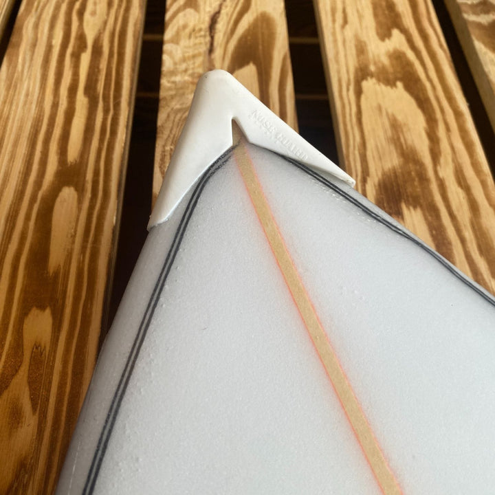 Eine Nahaufnahme zeigt die Nase eines weißen Surfbretts, das mit einem SurfCo Hawaii - Surfboard Noseguard ausgestattet ist und auf einer Holzoberfläche mit sichtbaren Maserungen ruht.