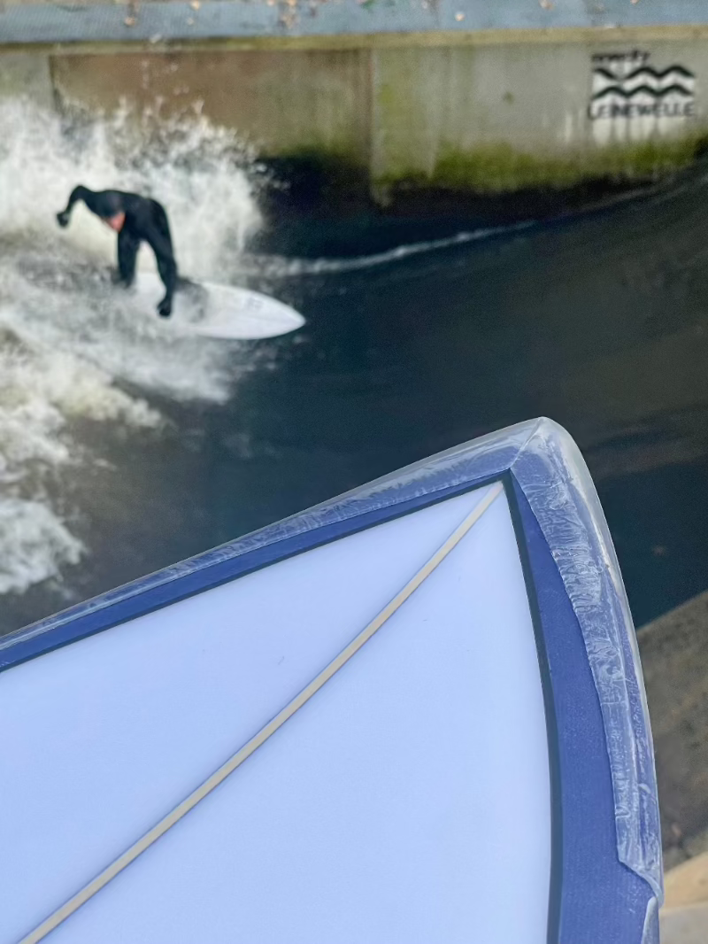 Eine Person in einem schwarzen Neoprenanzug surft auf einer künstlichen Welle in einer schmalen Wasserstraße aus Beton, wobei die Spitze eines SurfCo Hawaii - Surfboard Noseguard (Longboard) im Vordergrund zu sehen ist und die mit Graffiti beschmierten Wände im Hintergrund.