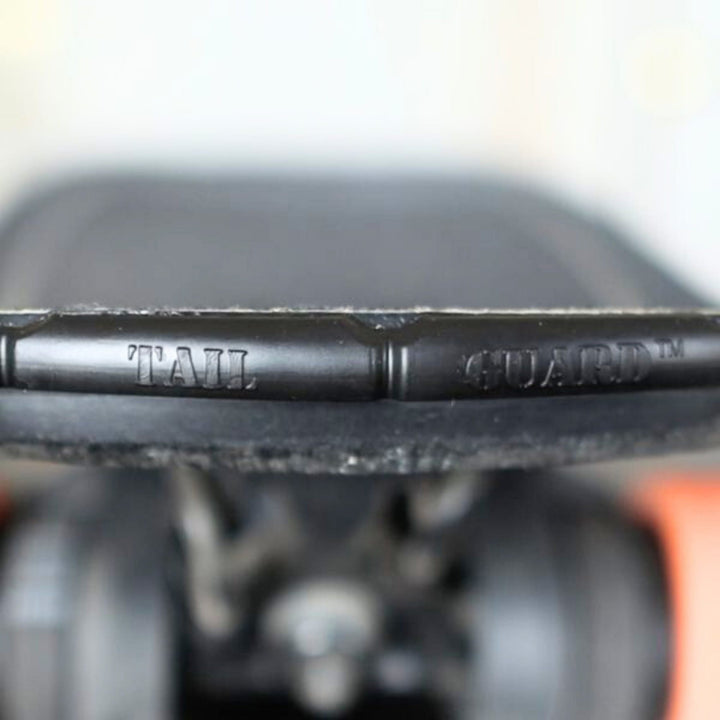 Nahaufnahme eines SurfCo Hawaii - Jumbo Tailguard von SurfCo Hawaii, das am Heck des Skateboards befestigt ist, mit eingraviertem Branding auf dem Schutzstreifen; Räder und Achsen sind darunter unscharf zu sehen.