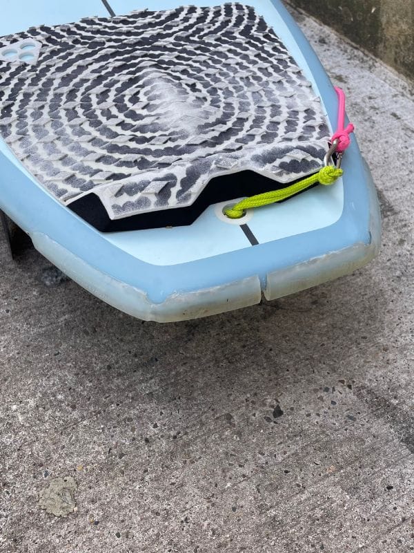 Nahaufnahme des hinteren Endes eines blauen Surfbretts, das auf Beton ruht. Zu sehen sind ein schwarz-weißes Traktionspad, eine gelbe Leine und einige sichtbare Abnutzungserscheinungen am Rand des Bretts.