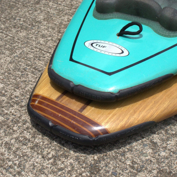 Nahaufnahme von zwei Paddleboard-Nasen auf Beton - eine in Türkis und Schwarz, die andere mit einem Holzmaserungsmuster - beide mit schwarzem Gummirand, ähnlich dem Schutz, den der SurfCo Hawaii - Surfboard Tailguard bietet.