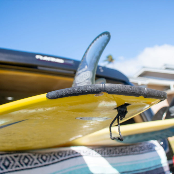 Nahaufnahme eines gelben Surfboards mit einem SurfCo Hawaii - Surfboard Tailguard, das auf einem gestreiften Handtuch in der Nähe eines Fahrzeugs ruht. Der klare blaue Himmel, Haus und Palme schaffen eine ideale Kulisse für jede SurfCo Hawaii Abenteuer.
