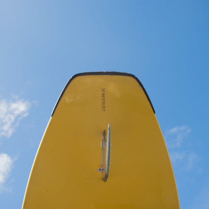 Eine Nahaufnahme des unteren hinteren Endes eines gelben Surfbretts mit einer einzigen Flosse und einem SurfCo Hawaii - Surfboard Tailguard vor einem blauen Himmel mit verstreuten Wolken.