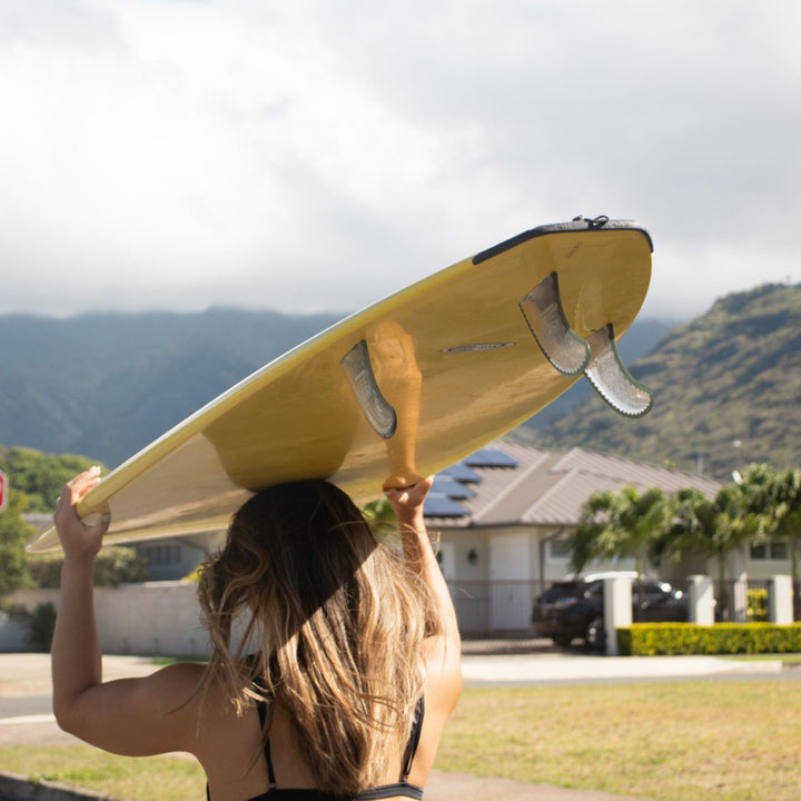 Eine Person mit langen Haaren trägt ein gelbes Surfbrett mit einem SurfCo Hawaii Tailguard über dem Kopf und geht an einem teilweise bewölkten Tag im Freien spazieren, mit Bergen, einem Haus und einem geparkten Auto im Hintergrund.