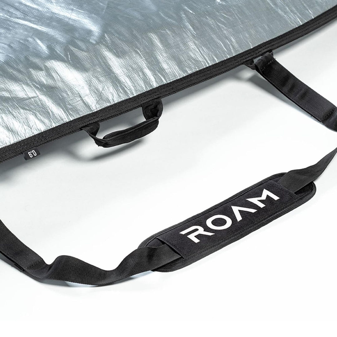 Eine Nahaufnahme eines schwarzen Griffs mit weißem ROAM-Branding auf einer strukturierten silbernen Roam Boardbag Surfboard Daylight Hybrid Fish, wobei auch zusätzliche schwarze Riemen sichtbar sind.