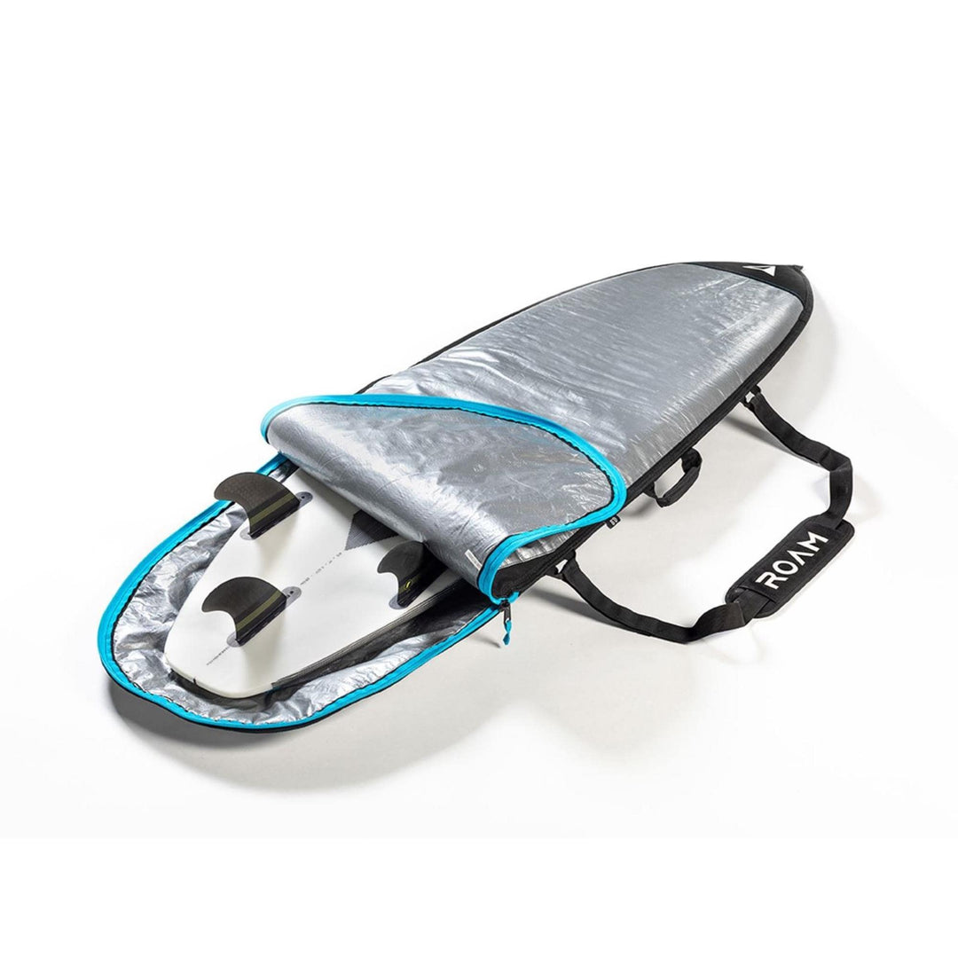 Eine silberne und blaue ROAM Boardbag Surfboard Daylight Shortboard ist teilweise geöffnet und zeigt ein dreiflügeliges Surfbrett im Inneren. Die Tasche hat einen schwarzen Schultergurt mit dem Aufdruck "RMW" auf dem Pad.