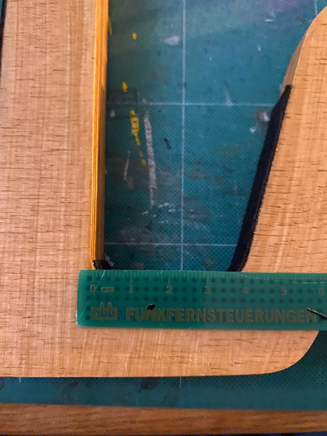 Ein grünes, transparentes Lineal mit der Aufschrift FUNKFERNSTEUERUNGEN misst eine Holzkante aus, möglicherweise für die Herstellung der Brandungsplanke Surfboard Wandhalterung aus Holz, auf einer blauen Schneidematte.