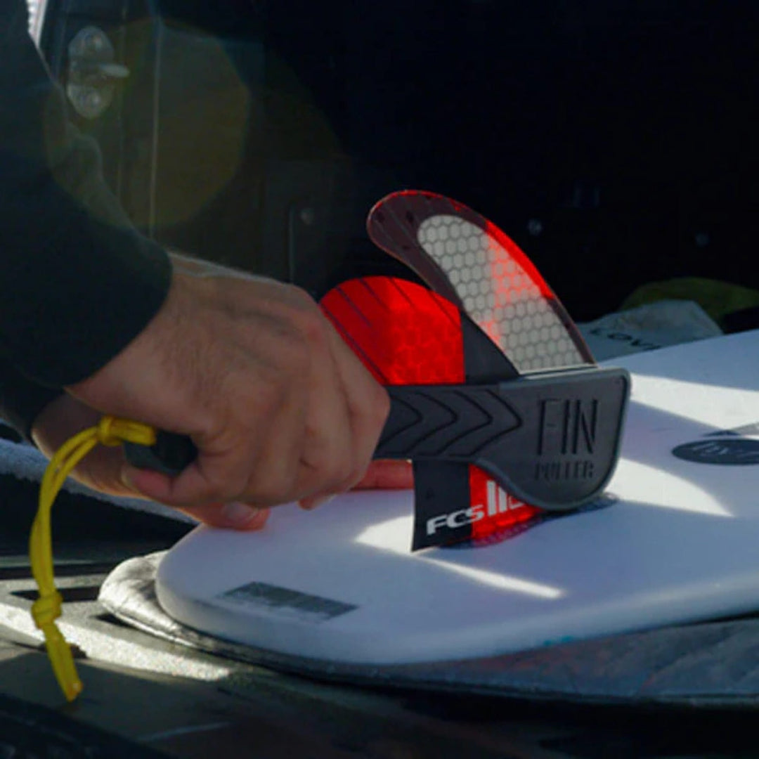 Eine Person benutzt den Finpuller für FCS2 Finnen von Finpuller, um eine rote Surfboardfinne auf einem weißen Brett im Freien, möglicherweise in der Nähe eines Fahrzeugs, einzustellen. Das Werkzeug bietet eine einfache Finnenentfernung für FCS2-Flossen.