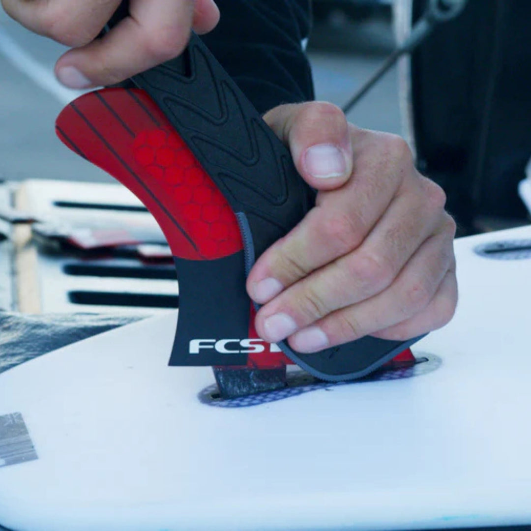 Eine Person benutzt beide Hände mit dem Finpuller für FCS2 Finnen von Finpuller, um eine FCS-Surfbrettfinne an einer weißen Tafel anzubringen oder zu entfernen, wobei die Außenumgebung im Hintergrund teilweise sichtbar ist.