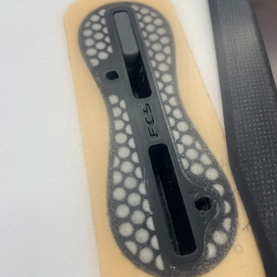 Nahaufnahme eines FCS II-Finnenkastens auf einem weißen Surfbrett mit Wabenboden, Finnenschraube und schwarzem Traktionspad; verwenden Sie das Rapid Surf Shop Tab Infill Kit, um ihn mit FCS I-Flossen kompatibel zu machen.