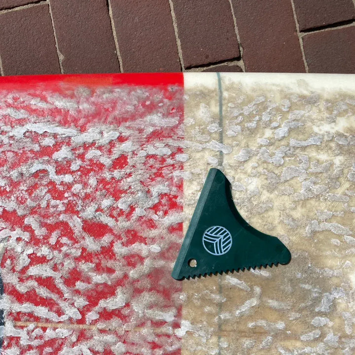 Nahaufnahme eines mit rotem und weißem Wachs überzogenen Surfbretts, mit einer Rebel Fin - Wachskamm von Sex Wax und Ziegelsteinpflaster im Hintergrund.