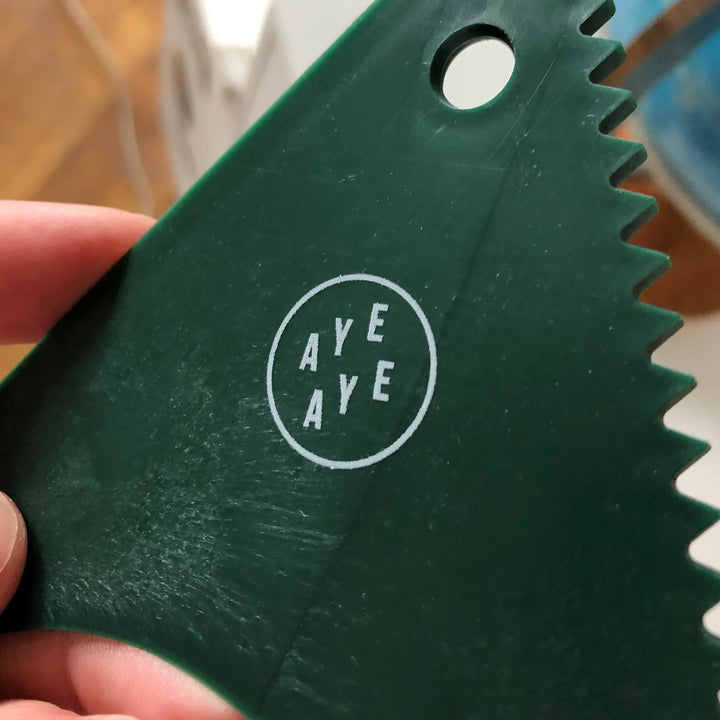 Eine Hand hält eine dunkelgrüne Rebel Fin - Wachskamm von Sex Wax, hergestellt aus recycelten Fischernetzen. Sie hat gezackte Kanten, ein rundes Loch in der Nähe der Ecke und zeigt ein weißes, rundes Logo mit "AYE AYE" in zwei Zeilen.