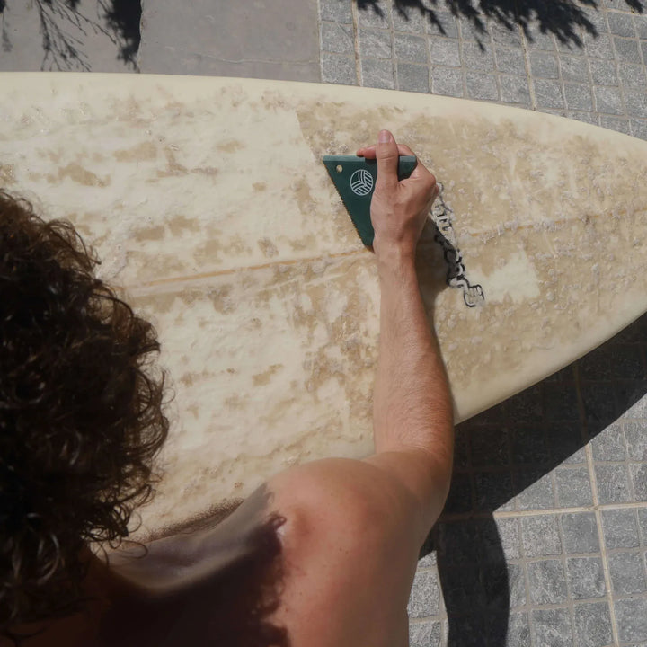 Eine Person benutzt eine Rebel Fin - Wachskamm von Sex Wax, um altes Wachs von einem Surfbrett abzukratzen, während sie im Sonnenlicht auf Fliesen im Freien steht.