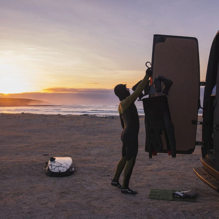 Bei Sonnenuntergang am Strand steht eine Person in einem Neoprenanzug an einer offenen Van-Tür und hält einen weiteren Neoprenanzug mit dem C-Monsta Wetsuit Aufhänger von C-Monsta. In der Nähe liegen ein Surfbrett und weiteres Wassersport-Zubehör auf dem Sand.