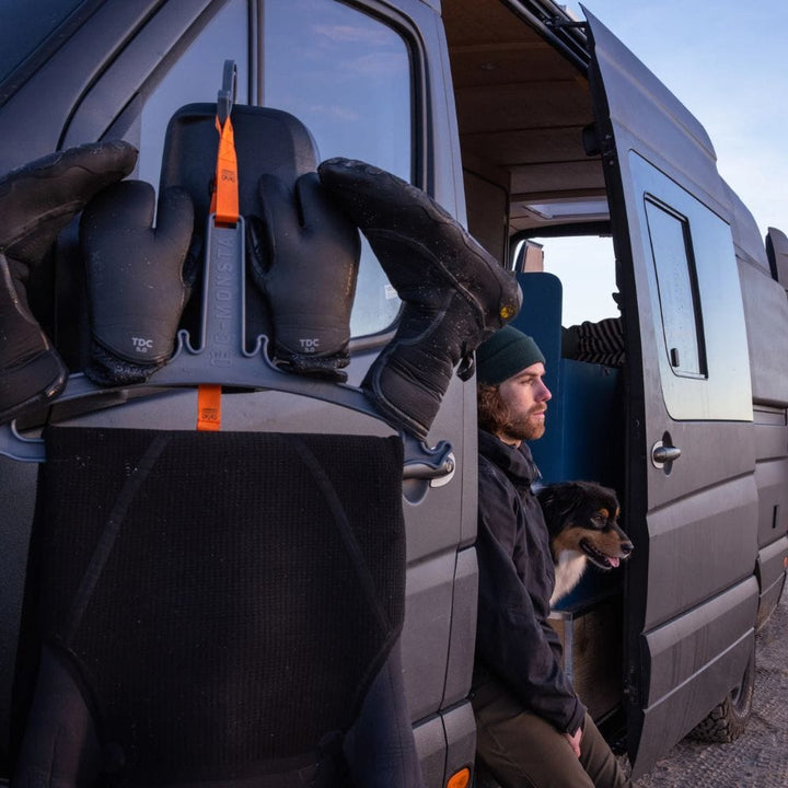 Ein Mann sitzt mit seinem schwarz-braunen Hund in der Tür eines dunklen Vans. Im Vordergrund hält C-Monsta Wetsuit Aufhänger von C-Monsta schwarze Handschuhe mit orangefarbenen Akzenten zwischen essentiellem Wassersport Zubehör.