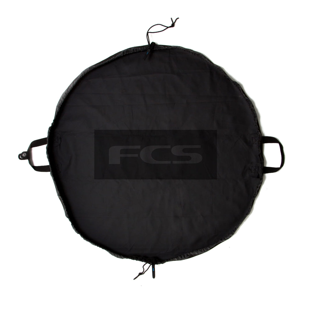 Die FCS - Change Mat ist eine runde, schwarze Matte mit zwei kurzen Griffen und einem Kordelzugverschluss. In der Mitte ist in großen grauen Buchstaben "FCS" aufgedruckt - ideal als praktischer Neoprenanzugbeutel.