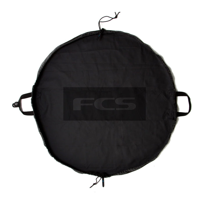 Die FCS - Change Mat ist eine runde, schwarze Matte mit zwei kurzen Griffen und einem Kordelzugverschluss. In der Mitte ist in großen grauen Buchstaben "FCS" aufgedruckt - ideal als praktischer Neoprenanzugbeutel.