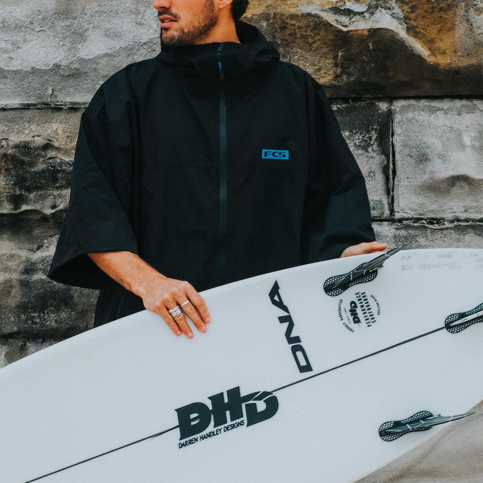 Eine Person in einem FCS All Weather Surf Poncho (wasserfest), schwarz, steht an einer Steinmauer und hält ein weißes Surfbrett mit FCS-Flossen und schwarzen Logos, darunter BHD und DNA.