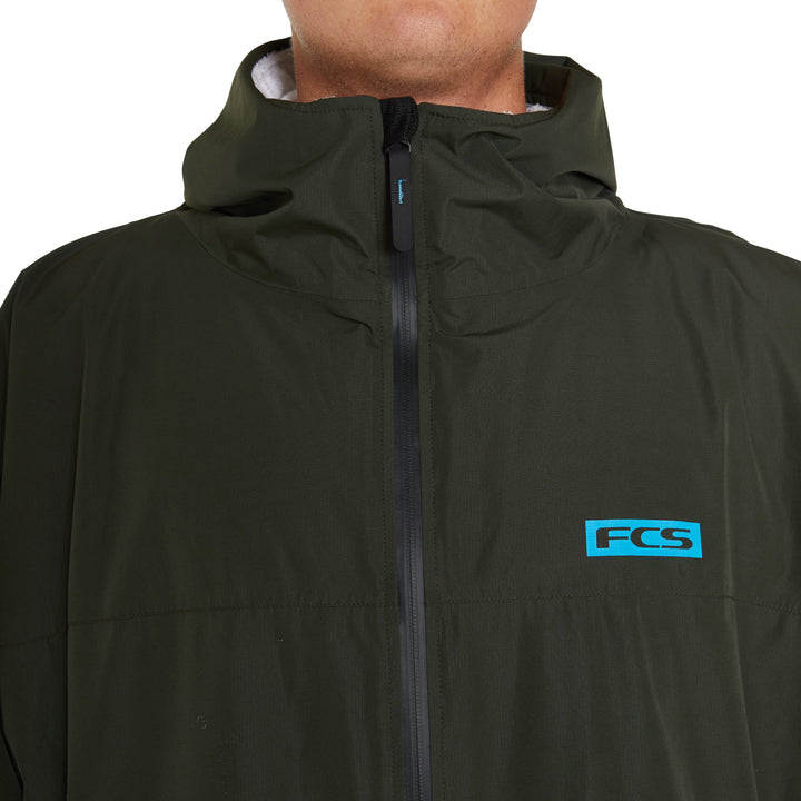 Eine Person, die einen dunkelgrünen FCS All Weather Surf Poncho (wasserfest) mit einem schwarzen Reißverschluss und einem blauen FCS-Logo auf der linken Brust trägt. Das Bild ist so beschnitten, dass nur das untere Gesicht, der Hals und der obere Brustbereich zu sehen sind.