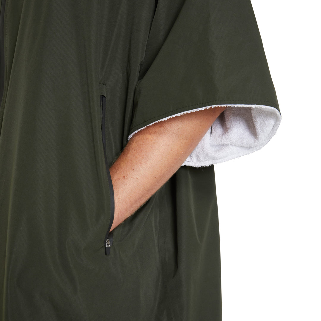Eine Person, die den FCS All Weather Surf Poncho (wasserfest) in Dunkelgrün mit weißem Futter trägt, steckt ihre Hand in eine Tasche mit Reißverschluss. Nur der Arm und der Oberkörper sind vor dem weißen Hintergrund zu sehen, was dieses FCS-Essential hervorhebt.
