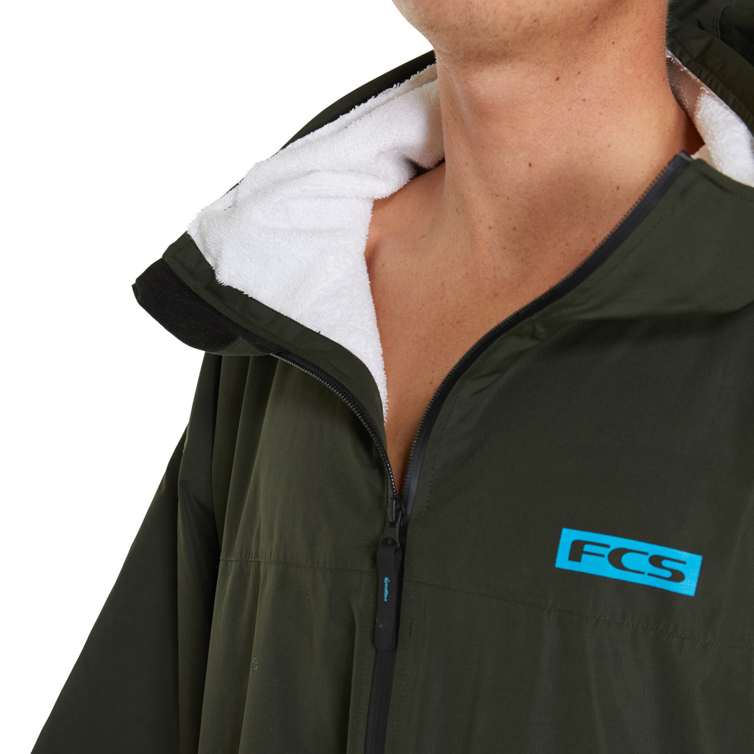Eine Person trägt den FCS All Weather Surf Poncho (wasserfest) - eine dunkelgrüne, wasserabweisende Jacke mit weißem Fleecefutter und einem blauen FCS-Logo auf der Brust. Das Gesicht ist größtenteils außerhalb des Rahmens. Perfekt zum Überziehen über den Surfponcho.