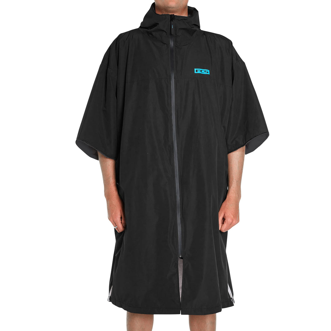 Eine Person, die den FCS All Weather Surf Poncho (wasserfest) in Schwarz mit kurzen Ärmeln und einem Frontreißverschluss trägt, steht vor einem schlichten weißen Hintergrund. Nur ihre Unterschenkel und Hände sind sichtbar.