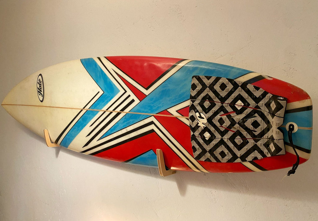 Ein Surfbrett mit einem roten, blauen und weißen geometrischen Muster wird mit der Brandungsplanke Surfboard Wandhalterung aus Holz an der Wand befestigt. Es verfügt über ein schwarz-weißes Trittbrett am Heck und ist mit einer Leine versehen.