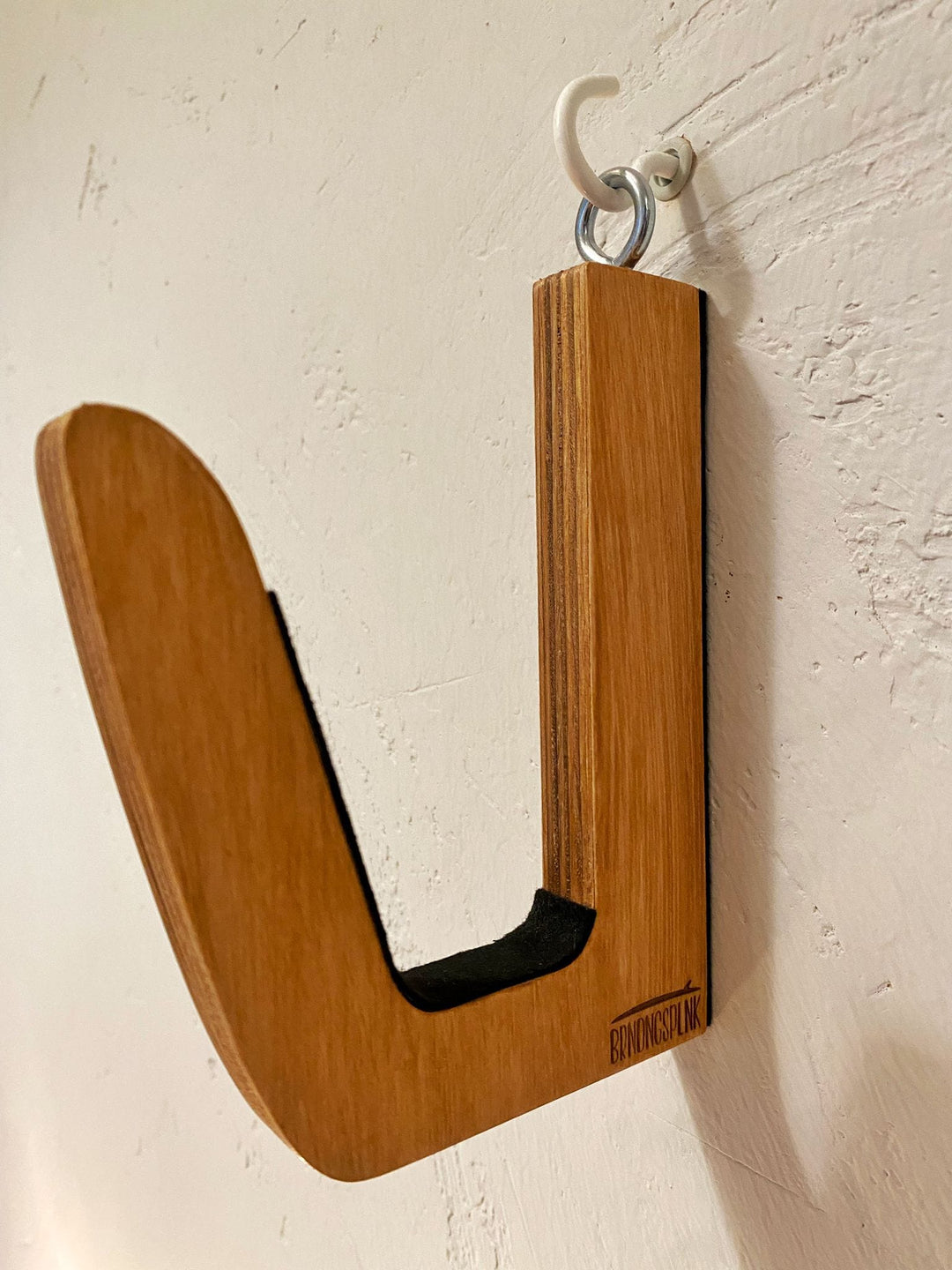 Die Brandungsplanke Surfboard Wandhalterung aus Holz ist eine hölzerne, J-förmige Surfboard-Wandhalterung mit natürlicher Oberfläche und schwarzer Polsterung, die mit einem Metallhaken sicher an einer weißen Wand befestigt wird.