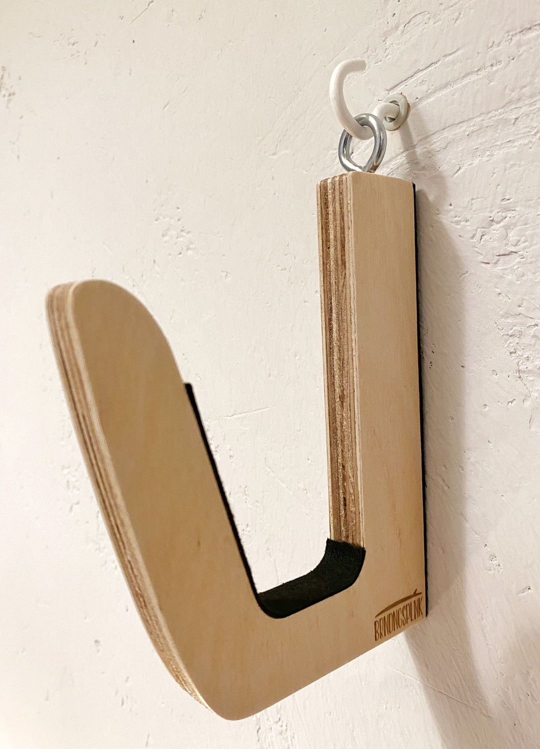 Eine Brandungsplanke Surfboard Wandhalterung aus Holz in J-Form ist mit einem weißen Schraubhaken an einer weißen Wand befestigt. Das geschichtete Holz erinnert an ein Surfbrett, ein kleines Logo befindet sich am rechten unteren Rand.