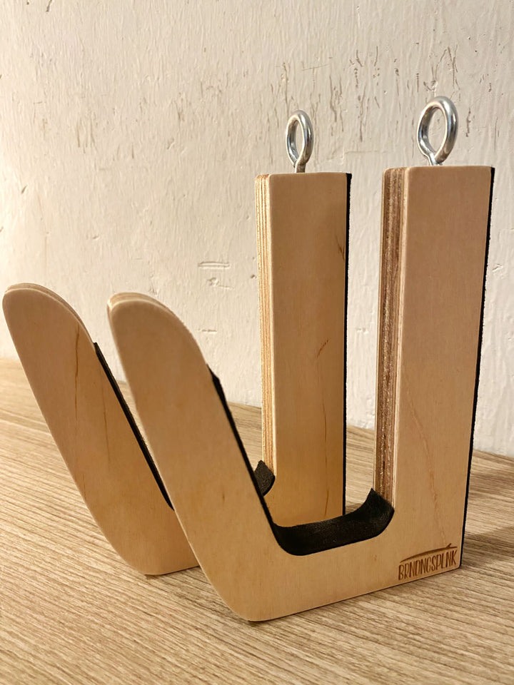 Die Surfboard Wandhalterung aus Holz von Brandungsplanke mit zwei gebogenen Armen und Metallösen steht auf einer Holzfläche vor einer strukturierten, cremefarbenen Wand.